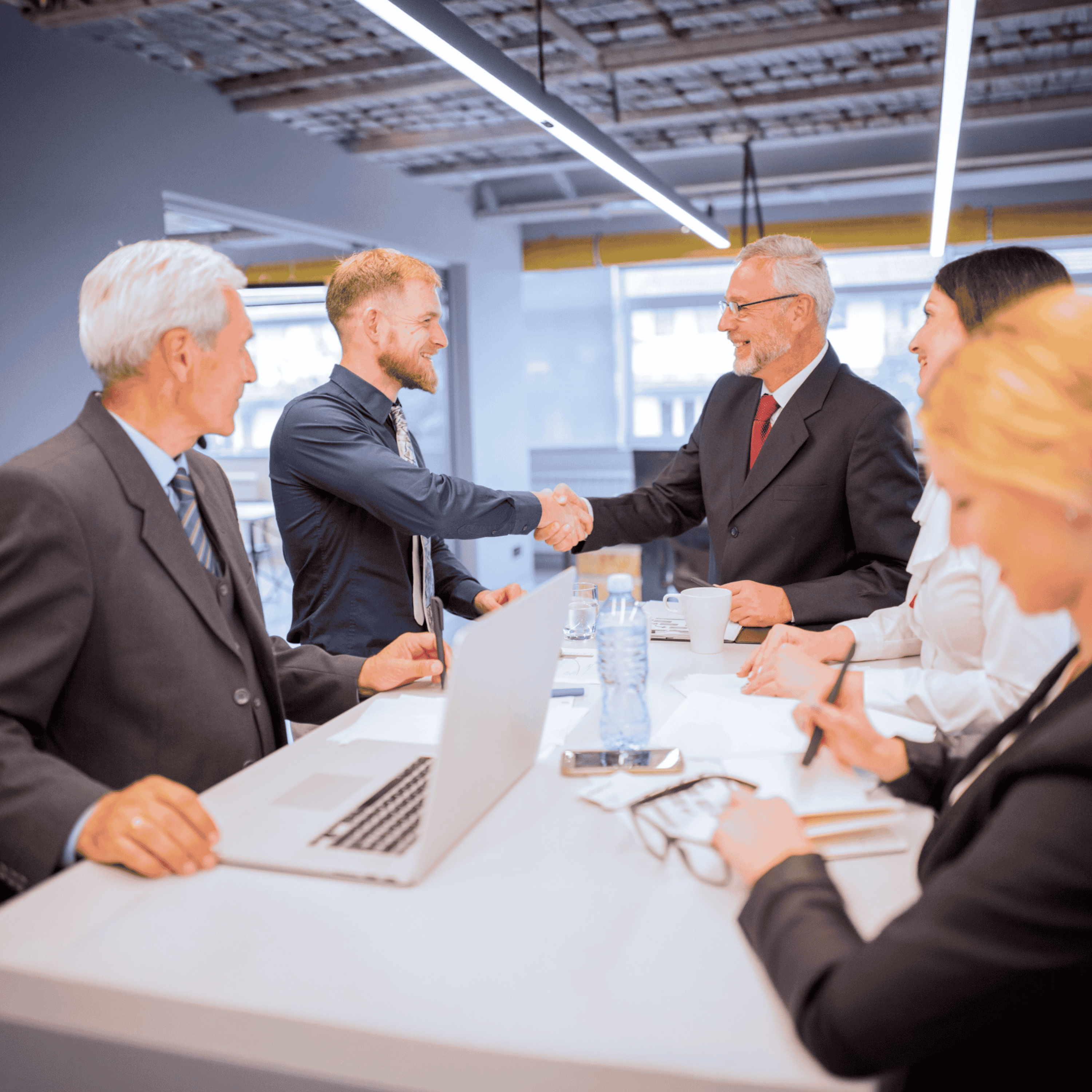 Five business professionals gather around a sleek white conference table in a modern office, where two men seal a deal with a firm handshake while colleagues observe with laptops, documents, and coffee cups spread across the table under bright overhead lighting.